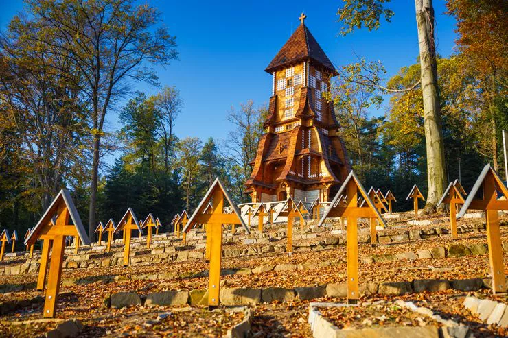 Ansicht des Hügels von unten mit den sichtbaren Grabstätten. Über jeder der Grabstätten steht ein überdachtes Holzkreuz. Dahinter ist ein hoher Holzturm zu sehen - eine Schindel und im Hintergrund ein Herbstwald.