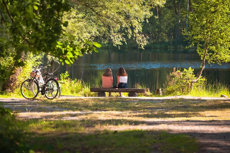 Rückansicht von zwei Mädchen, die an einem sonnigen Tag auf einer Holzbank sitzen und auf die Oberfläche des Sees schauen.  Neben ihnen sind zwei Fahrräder aufgestellt. Ringsum sind grüne Bäume und ein Waldweg zu sehen.