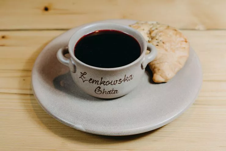 A cup filled with red borscht is standing on a saucer next to a peroh dough pocket with savoury filling. The whole is standing on a white wooden table.