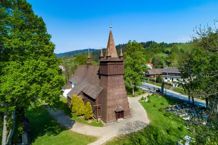 A drone-eye view of a wooden church surrounded by greenery and trees. To the right, the grave stones of a small churchyard can be seen, and beyond them a road and some houses.
