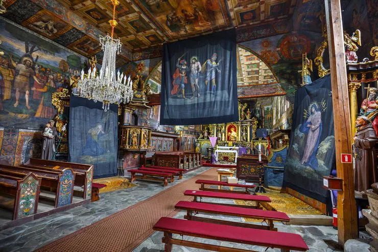 Blick auf das Innere der Holzkirche. An den Wänden sind zahlreiche Gemälde zu sehen, in der Mitte stehen Kirchenbänke. An den beiden Seitenaltären und in der Mitte hängen große Tücher, auf denen Szenen aus dem Kreuzweg dargestellt sind.