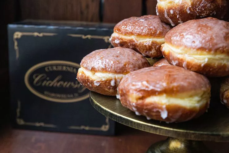 A pyramid of round deep-fried and lavishly glazed doughnuts is standing on a metal tray in front of an elegant box bearing the logo of Cichowscy Confectionery.