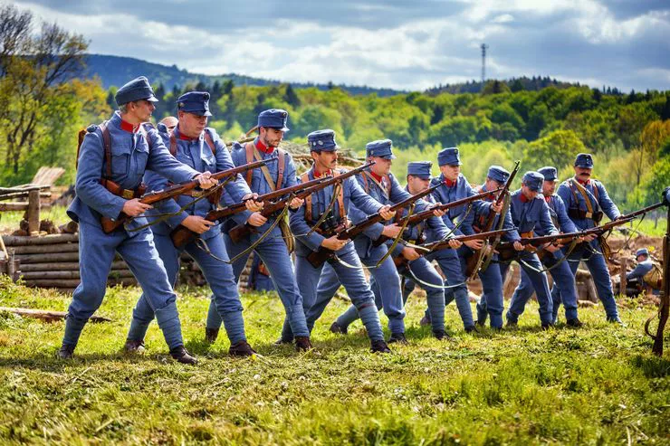 A line of men wearing blue military uniforms hold their position in a sun-drenched meadow. Each is holding a rifle ready to fire. The background consists of green hills.